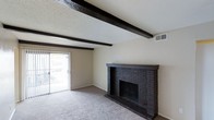 This single-floor space offers a welcoming and characterful living environment defined by a thoughtful blend of rustic and classic design elements. The main living area immediately draws the eye upward to a textured ceiling accented by striking dark-stained exposed wood beams that span the width of the room, adding architectural depth and a warm, timeless character. Anchoring the space is a substantial floor-to-ceiling brick fireplace finished in a dark tone, serving as a bold focal point that lends both visual drama and functional warmth to the room. The neutral, wall-to-wall carpeting throughout provides a soft, comfortable foundation that complements the darker accents above. Large sliding glass doors along one wall invite an abundance of natural light, brightening the interior and providing a seamless visual connection to the outdoor area beyond. The open layout of the living space feels generously proportioned, offering flexibility for a variety of furniture arrangements. The combination of the beamed ceiling, brick fireplace, and light-filled atmosphere creates an inviting ambiance that balances coziness with an open, airy feel throughout the home.