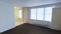 This single-floor space presents a straightforward, functional layout characterized by a combination of practical finishes and natural light. The main living area features dark wall-to-wall carpeting that contrasts with the light yellow-painted walls, creating a neutral yet warm atmosphere. A set of large windows fitted with horizontal blinds lines one wall, allowing generous amounts of natural light to fill the room while also housing a baseboard heating unit beneath them. A supporting column is visible near the transition between spaces, adding a subtle architectural element to the open layout. An adjoining area, visible through the open passage, continues the yellow wall color scheme and includes an additional window, further enhancing the overall brightness of the space. The ceiling is smooth and white, complemented by a flush-mounted light fixture. The open flow between the two areas provides a sense of connected, usable space, while the minimal design keeps the focus on functionality. The combination of natural light, neutral tones, and practical heating infrastructure makes this space a clean, move-in ready environment suited for everyday living.