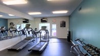 This single-floor fitness center is designed to provide a functional and comfortable workout environment. The space is outfitted with a selection of cardio equipment, including treadmills and an elliptical machine, positioned to make efficient use of the available floor area. Dark rubber flooring covers the ground, offering durability and safety during exercise.

A full-length mirror runs along one wall, enhancing the sense of space while allowing users to monitor their form. Multiple wall-mounted flat-screen TVs are positioned for easy viewing from the cardio equipment, adding an entertainment element to workouts. Overhead fluorescent lighting provides consistent, even illumination throughout the room.

The walls feature a neutral white finish complemented by a teal accent wall, giving the space a clean and modern aesthetic. A posted fitness center rules board is displayed near the entrance, and a sanitation station is conveniently located nearby. Natural light filters in through windows and a set of glass-panel double doors that lead directly to the exterior, contributing to an open and airy atmosphere. The textured ceiling and overall layout reflect a practical, well-maintained facility suited for everyday fitness use.
