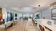 Located at 1025 Hirschfeld Dr in Fayetteville, NC, this expansive single-story community clubhouse offers a generously sized, thoughtfully designed gathering space for residents. The open-concept layout is anchored by a bright, airy leasing lounge featuring an arched entryway, large windows that draw in abundant natural light, light wood-style flooring, and elevated ceilings with decorative tray details and herringbone wood inlays. A central reception desk sits atop a patterned area rug, framed by botanical-print accent walls and curated artwork. The lounge flows seamlessly into a spacious social area furnished with comfortable seating, oversized circular pendant lighting, and views of the surrounding outdoor areas through full-height glass partition walls. The kitchen and bar area is a standout feature, showcasing a large white quartz island with bar seating, dark cabinetry with a striking herringbone tile backsplash, and modern globe pendant lighting. The fitness wing includes a well-appointed gym with cardio machines, free weights, and full-wall mirrors, complemented by a dedicated yoga and cycling studio with natural light and outdoor views. A café-style nook adorned with framed landscape artwork rounds out this polished, amenity-rich community hub.