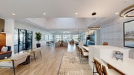 Located at 1025 Hirschfeld Dr in Fayetteville, NC, this expansive single-story community clubhouse offers a generously sized, thoughtfully designed gathering space for residents. The open-concept layout is anchored by a bright, airy leasing lounge featuring an arched entryway, large windows that draw in abundant natural light, light wood-style flooring, and elevated ceilings with decorative tray details and herringbone wood inlays. A central reception desk sits atop a patterned area rug, framed by botanical-print accent walls and curated artwork. The lounge flows seamlessly into a spacious social area furnished with comfortable seating, oversized circular pendant lighting, and views of the surrounding outdoor areas through full-height glass partition walls. The kitchen and bar area is a standout feature, showcasing a large white quartz island with bar seating, dark cabinetry with a striking herringbone tile backsplash, and modern globe pendant lighting. The fitness wing includes a well-appointed gym with cardio machines, free weights, and full-wall mirrors, complemented by a dedicated yoga and cycling studio with natural light and outdoor views. A café-style nook adorned with framed landscape artwork rounds out this polished, amenity-rich community hub.