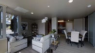 This single-floor space showcases a thoughtfully designed open-concept layout that seamlessly connects the living, dining, and kitchen areas into one cohesive environment. The living area is anchored by a light-toned sofa accented with decorative pillows, complemented by a sleek arc floor lamp with multiple shades that provides layered ambient lighting. A low-profile side table with a metal frame and wood shelving adds functional storage alongside the seating area. The space features light hardwood-style flooring that runs throughout, contributing to a clean, modern aesthetic. Recessed ceiling lighting enhances the contemporary feel, while a decorative art piece on the wall adds a pop of personality. The dining area, positioned adjacent to the living space, includes a glass-top table surrounded by upholstered side chairs. The kitchen, visible in the background, is fitted with dark cabinetry, light countertops, and stainless steel appliances, offering a refined contrast to the neutral palette of the surrounding space. A structural column defines the transition between zones without fully enclosing any area, preserving the open flow. Overall, the space presents a modern, urban ambiance with functional, well-considered design choices.