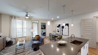 This single-floor residence is designed with a cohesive, modern aesthetic that balances functionality and style throughout. The entry opens adjacent to a well-equipped kitchen featuring white shaker cabinetry, stainless steel appliances, a hexagonal tile backsplash, and a large island topped with dark stone countertops. Pendant lighting illuminates the island, which also houses an undermount sink. In-unit laundry is conveniently located nearby.

The open-concept layout connects the kitchen seamlessly to a dining area and living room. The dining space accommodates a full table and chairs beneath additional pendant lighting, while bold artwork anchors the wall behind it. The living room features hardwood-style flooring, a ceiling fan, large windows with decorative curtains that invite natural light, and ample space for comfortable seating.

The bedroom is a calm retreat with soft carpet, a ceiling fan, natural light from a window, and a neutral accent wall displaying framed artwork. Two bathrooms serve the unit: one with a soaking tub, white subway tile, and built-in cabinetry; the other with a glass-enclosed shower, extended vanity with dark stone countertops, and access to a walk-in closet with organized shelving.