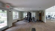 This fitness room is a well-equipped, single-floor exercise space designed to support a variety of workout routines. The room is anchored by a large multi-station cable machine at its center, which accommodates several exercise configurations simultaneously. Flanking the cable system are additional strength-training stations, including a pull-up and dip tower, along with free weights and benches positioned throughout the space. Cardio equipment is visible toward the rear of the room, providing options for aerobic training.

The flooring consists of patterned carpet tiles in earthy tones, offering cushioning underfoot while defining the workout area. Recessed lighting is evenly distributed across the textured white ceiling, ensuring consistent illumination throughout. Large windows along one wall invite natural light into the room and provide views of the surrounding brick exterior, contributing to an open, airy feel. The walls are finished in a neutral white, keeping the environment clean and straightforward. Overall, the space is functional and practical, offering residents or users a dedicated area for comprehensive fitness training.
