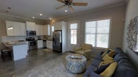 This single-story home offers a seamlessly integrated open-concept layout that connects the living area and kitchen into one cohesive, functional space. The kitchen is outfitted with stainless steel appliances, including a French door refrigerator, a gas range, a microwave, and a dishwasher, all complemented by crisp white cabinetry and light countertops. Recessed lighting throughout the kitchen ensures excellent illumination, while crown molding along the ceiling adds a refined architectural touch.

The living area is warm and inviting, anchored by a ceiling fan with a light fixture that enhances airflow and comfort. Large windows fitted with white horizontal blinds allow generous natural light to fill the space, creating a bright and airy atmosphere. A glass-paned exterior door further connects the interior to the outdoors, reinforcing the open feel of the home.

The flooring consists of polished concrete, lending the space an understated, modern aesthetic that is both durable and easy to maintain. An area rug defines the living zone, which comfortably accommodates a sofa, an accent chair, and a patterned upholstered ottoman that serves as a central focal point. The neutral color palette throughout—warm grays, whites, and soft yellow accents—contributes to a cohesive and welcoming ambiance.