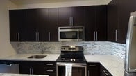 This single-floor space showcases a thoughtfully designed interior with a strong emphasis on modern aesthetics. The kitchen stands as a centerpiece, featuring rich, dark espresso-toned cabinetry that extends from floor to ceiling, maximizing storage while creating a bold visual statement. Stainless steel appliances, including an over-the-range microwave and a freestanding electric range, lend a polished, professional quality to the space. The light-toned stone countertops provide a striking contrast against the dark cabinetry, offering ample preparation and workspace. A stainless steel undermount sink with a contemporary gooseneck faucet adds both functionality and style. The kitchen backsplash features a linear mosaic tile pattern in neutral tones of white, grey, and silver, adding texture and visual interest to the walls. The clean lines of the flat-panel cabinet doors, accented by slim bar-style hardware, reinforce the contemporary design throughout. The overall ambiance of the space is sleek and cohesive, blending dark and light elements to create a sophisticated, modern environment well-suited for everyday living.