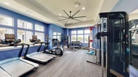 Located in Pryor, Oklahoma, this fitness center offers a comprehensively outfitted exercise space designed to support a wide range of workout routines. The room is defined by bold blue accent walls, a tray ceiling with recessed lighting, and a large industrial-style ceiling fan that ensures comfortable air circulation throughout. Durable patterned carpet flooring covers the entire space, providing traction and cushioning underfoot.

Natural light streams in through multiple large windows along the walls, offering views of the surrounding outdoor area, including a glimpse of a pool. Full-length mirrors are strategically mounted on one wall, visually expanding the space and allowing users to monitor their form during exercise.

The cardio zone is equipped with treadmills, an elliptical trainer, and a stationary bike. Strength training is well-supported with a multi-tiered dumbbell rack, a weight bench, and a cable machine. A prominent Octagon combat training station serves as a centerpiece, complete with heavy punching bags and resistance rope attachments. A medicine ball and kettlebell rack round out the functional fitness offerings, alongside colorful stability balls for core training. The space is thoughtfully organized to accommodate multiple users engaged in different workout modalities simultaneously.