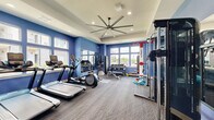 Located in Pryor, Oklahoma, this fitness center offers a comprehensively outfitted exercise space designed to support a wide range of workout routines. The room is defined by bold blue accent walls, a tray ceiling with recessed lighting, and a large industrial-style ceiling fan that ensures comfortable air circulation throughout. Durable patterned carpet flooring covers the entire space, providing traction and cushioning underfoot.

Natural light streams in through multiple large windows along the walls, offering views of the surrounding outdoor area, including a glimpse of a pool. Full-length mirrors are strategically mounted on one wall, visually expanding the space and allowing users to monitor their form during exercise.

The cardio zone is equipped with treadmills, an elliptical trainer, and a stationary bike. Strength training is well-supported with a multi-tiered dumbbell rack, a weight bench, and a cable machine. A prominent Octagon combat training station serves as a centerpiece, complete with heavy punching bags and resistance rope attachments. A medicine ball and kettlebell rack round out the functional fitness offerings, alongside colorful stability balls for core training. The space is thoughtfully organized to accommodate multiple users engaged in different workout modalities simultaneously.