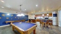 This single-floor amenity and recreation space offers a well-appointed layout designed for social gatherings and leisure activities. The room is anchored by a full-size billiards table with a vivid blue felt surface, positioned beneath a pendant light fixture, with a mounted flat-screen television and wall-mounted cue rack nearby. A shuffleboard table lines one wall, adding to the recreational offerings.

The fully equipped kitchen is a standout feature, showcasing warm-toned wood cabinetry, granite countertops, a large central island with a cooktop, stainless steel appliances including a double wall oven and French door refrigerator, and a tile backsplash. Pendant lighting illuminates the workspace, while slate-style tile flooring runs throughout the entire space, providing a cohesive, durable foundation.

A bold cobalt blue accent wall adorned with decorative sunburst medallions adds a vibrant visual element to the lounge area. High-top tables with bar stools provide casual seating, complemented by a round table grouping near glass entry doors that invite natural light. Recessed lighting throughout ensures the space remains bright and functional. The overall ambiance is lively yet practical, suited for both organized events and informal social use.