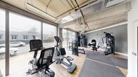 This fitness center in Miami's Brickell area offers a thoughtfully designed exercise space with an industrial-modern aesthetic. The room features exposed concrete ceilings with visible ductwork and piping, lending an urban, loft-style character throughout. Light wood-tone flooring contrasts with the raw concrete walls and gray painted surfaces, creating a clean and functional environment.

Large floor-to-ceiling windows and sliding glass doors line one side of the space, flooding the interior with natural light and providing street-level views of the surrounding neighborhood. The open layout accommodates a comprehensive selection of fitness equipment, including cardio machines such as treadmills, ellipticals, and a stationary bike, alongside a robust array of strength-training equipment including cable machines, Nautilus weight machines, a multi-station functional trainer, adjustable benches, and free weights. Protective rubber floor mats are positioned throughout the workout zones for safety and comfort.

A dedicated mirrored area supports stretching and floor exercises, while a small enclosed utility room provides additional infrastructure support. Strip lighting mounted to the ceiling ensures even, practical illumination across the entire floor. The space is well-suited for a full-body workout routine, offering both cardiovascular and resistance training options in a compact yet efficiently organized setting.