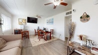 This single-story home is designed with a practical, open layout that connects the main living spaces efficiently. The living area is bright and welcoming, featuring wood-look laminate flooring, a ceiling fan, and windows that allow natural light to fill the room. A sofa, whiteboard, and decorative accents give the space a relaxed yet functional character, with an open sightline into the kitchen beyond.

The kitchen has been updated with white shaker-style cabinetry, a white subway tile backsplash, light stone-look countertops, a double-basin sink with a matte black faucet, and a stainless steel gas range with an overhead ventilation hood—a clean, modern aesthetic throughout.

A dedicated office or multipurpose area is set apart from the main living space, featuring exposed painted block walls, wood-look flooring, a wall-mounted flat-screen television, and ample desk and table space suited for work or study.

The home includes multiple bedrooms, each finished with the same wood-look flooring. One bedroom features decorative circular wall art and soft pink accents; another is furnished with a full-size bed framed by a window; a third accommodates a full-over-full metal bunk bed. The bathroom is clean and straightforward, offering a tub-shower combination, toilet, and vanity with tile-look flooring.