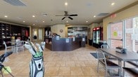 This single-floor golf facility clubhouse serves as a central hub for golfers, combining a well-equipped pro shop, a food and beverage counter, and a comfortable lounge area into one cohesive space. The interior is finished with light stone-look tile flooring throughout, lending a clean and consistent aesthetic across all areas.

The pro shop section showcases golf bags, apparel, and accessories, with display racks and shelving units housing a range of merchandise. Large windows along the perimeter flood the retail area with natural light while offering views of the surrounding greenery.

Adjacent to the retail space, a wrap-around service counter with a dark laminate finish serves as both a snack bar and check-in station. Behind the counter, a commercial kitchen area is visible, equipped with stainless steel appliances and ventilation. A beverage refrigerator, coffee station, and packaged snack displays make this a convenient stop for golfers before or after a round.

The lounge and seating area features metal café-style tables and chairs arranged beneath a ceiling fan, creating a relaxed gathering spot. Wall-mounted televisions provide entertainment, while multiple large windows and glass doors invite natural light and offer views of the course exterior. The overall ambiance is functional yet welcoming, designed to serve the practical needs of an active golf clientele.