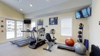 This single-floor fitness room offers a practical and well-organized space for exercise, outfitted with a variety of cardio and strength equipment. The room features treadmills, an elliptical trainer, and a recumbent stationary bike, providing options for users of varying fitness levels and preferences. Exercise balls and a step platform are also available, supporting flexibility, balance, and low-impact training routines.

The walls are painted in a warm neutral tone, contributing to an inviting atmosphere. Motivational fitness artwork is displayed prominently, adding an energetic visual element to the space. Two wall-mounted flat-screen televisions are positioned for easy viewing during workouts, enhancing the overall exercise experience.

Natural light enters through multiple windows fitted with white horizontal blinds, keeping the space bright while allowing for privacy and glare control. Recessed ceiling lighting supplements the natural light, ensuring the room remains well-lit at all times. The flooring is a durable, textured carpet suited for fitness use. Overall, the space is thoughtfully arranged to maximize functionality while maintaining a clean and welcoming environment.