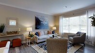 This single-floor space centers around a welcoming living room designed with a clean, contemporary aesthetic. The room features hardwood-style flooring that adds warmth and continuity underfoot, complemented by neutral beige walls that create a calm, inviting atmosphere. A large window dressed with white blinds and sheer curtains allows abundant natural light to fill the space, lending an airy, open quality to the interior.

The living area is thoughtfully arranged, anchored by a light-toned sofa accented with navy and coral throw pillows, paired with complementary seating and a textured area rug featuring a classic blue-and-cream pattern. A large landscape painting in muted blues and grays serves as a striking focal point on the main wall, adding a touch of artistic character. Flanking table lamps provide soft, ambient lighting throughout the room.

Side tables and a central coffee table offer functional surfaces without overcrowding the space, maintaining an open, uncluttered feel. The overall ambiance is relaxed yet polished, blending comfort with tasteful design details that make the living area both practical and visually appealing.