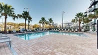 Located at 3701 SW 34th St in Gainesville, Florida, this outdoor amenity area offers a resort-inspired retreat designed for relaxation and recreation. At the heart of the space is a generously sized swimming pool with crystal-clear blue water, accented by a built-in waterfall feature that adds both visual interest and ambient sound. The pool is complemented by a broad, paver-tiled sun deck that provides ample room for lounging and socializing.

Surrounding the pool, rows of modern white chaise lounge chairs are thoughtfully arranged to capture Florida's abundant sunshine. Distinctive sculptural shade canopies are positioned throughout the deck, offering stylish relief from the sun. Tall, mature palm trees are interspersed throughout the landscaped areas, reinforcing the tropical atmosphere and providing natural shade.

Lush plantings of ornamental grasses, shrubs, and ground cover line the perimeter, softening the hardscape and adding vibrant greenery to the environment. A sleek black iron fence encloses the entire area, ensuring privacy and security while maintaining open sightlines to the sky.

The multi-story residential building rises directly adjacent to the pool area, with private balconies overlooking the deck below. The overall design blends contemporary architecture with a warm, leisure-focused outdoor environment, making this amenity space a standout feature of the property.