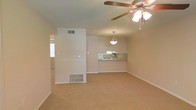 This single-floor space features an open-concept layout that connects the living area to the kitchen, creating a cohesive and functional floor plan. The living area is finished with wall-to-wall carpet in a neutral beige tone, complemented by clean, light-colored walls that lend a bright and airy feel to the interior. A ceiling fan with an integrated light fixture is centrally mounted, providing both ventilation and ambient lighting. A floor vent is visible along the baseboard, indicating a forced-air HVAC system for climate control. The kitchen is accessible via a pass-through counter with mirrored or reflective tile backsplash detailing, adding a subtle visual element to the space. A pendant light hangs above the counter, illuminating the transitional area between the kitchen and living room. A doorway to the left leads to an additional room or corridor, while a window on the far left allows natural light to enter. The overall ambiance is clean, neutral, and move-in ready, suited for straightforward, comfortable everyday living.
