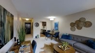 This single-floor residence offers an open-concept layout that integrates the living, dining, and kitchen areas into one cohesive and functional space. The living area is anchored by a contemporary sofa accented with bold blue throw pillows, complemented by a round wooden coffee table and a cowhide area rug that adds texture and warmth to the light-toned flooring. Decorative wall accents and framed artwork contribute to the space's curated aesthetic.

The dining area transitions naturally from the living space and features a round dining table surrounded by navy blue upholstered chairs, creating an inviting setting for meals. A flush-mount ceiling light overhead provides focused illumination for the dining experience.

The kitchen is visible beyond the dining area and is outfitted with dark cabinetry, providing a strong contrast against the lighter walls and countertops. It is efficiently designed to maximize functionality within a compact footprint.

The entryway, visible at the far end of the space, features a dark front door and is accented with wall décor including a large round clock, setting a stylish first impression. Throughout, neutral wall tones and consistent lighting create a bright, welcoming ambiance that ties all areas together harmoniously.