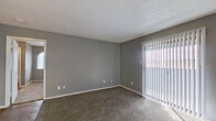 This single-floor space presents a cohesive, neutral aesthetic throughout, with gray walls and white trim creating a clean, modern backdrop across all rooms. The layout, as revealed by the overhead floor plan view, includes a living area, multiple bedrooms, a kitchen, and a bathroom, all arranged in a functional configuration.

The living area is generously sized, finished with wall-to-wall carpet and featuring a large sliding glass door dressed with vertical blinds, allowing ample natural light to fill the space. A textured ceiling adds subtle architectural detail throughout the unit.

The bedrooms continue the carpeted flooring and gray color palette. Each room is equipped with a ceiling fan and light fixture, and benefits from natural light through windows, including one room with a distinctive arched window. One bedroom connects directly to a bathroom featuring marble-style tiled shower walls and hard surface flooring.

The kitchen stands out with rich dark-wood cabinetry, light granite-style countertops, a mosaic tile backsplash, black appliances, and wood-look flooring—offering a sharp contrast to the softer tones found throughout the rest of the space.

An entry area transitions between the hard-surface flooring of the kitchen and the carpeted living zones, completing this well-organized, move-in ready layout.