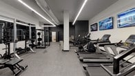 This fitness facility is designed to support a comprehensive workout experience within a single-floor layout. The space features a clean, modern aesthetic with crisp white and dark gray walls, rubberized flooring throughout, and bright linear LED lighting that runs across the ceiling to provide consistent illumination across the entire area.

The main workout floor is generously sized and thoughtfully organized. The cardio section includes multiple Spirit treadmills, an air bike, and additional cardio equipment, all positioned alongside a wall-mounted row of flat-screen televisions for entertainment during workouts. A dedicated strength training area houses a power rack with weight plates, multiple adjustable benches, a full dumbbell rack, medicine balls, and a wall-mounted pull-up bar — offering a complete range of resistance training options.

Structural columns are integrated into the layout, defining different workout zones without enclosing them, which maintains an open and airy feel. A staircase with modern cable railings and dual handrails provides access to an upper entry point. Exposed ductwork along the ceiling adds an industrial design element that complements the facility's contemporary aesthetic.

At the rear of the space, a darkened corridor leads to dedicated restroom facilities, including separate gendered bathrooms and a shower room, ensuring full convenience for users before and after their workouts.