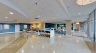This sophisticated single-floor commercial space is designed to impress, offering a seamless blend of professional functionality and contemporary aesthetics. The grand entrance features polished marble tile flooring and floor-to-ceiling glass walls and doors that flood the interior with natural light while providing views of lush outdoor greenery.

The expansive main lobby serves as the heart of the space, showcasing a gleaming marble floor, recessed lighting, and a drop ceiling with a clean, grid-panel design. Bold teal accent walls punctuate the open layout, adding a vibrant yet professional color palette throughout. Multiple distinct lounge areas are thoughtfully arranged, each furnished with modern sofas, armchairs, area rugs, and accent tables — ideal for informal meetings or relaxed waiting.

A sleek reception area features a prominent desk, a mounted flat-screen display, and warm lighting, offering a welcoming point of contact. A central high-top bar table with bar stools provides a casual collaborative zone. Additional seating alcoves along the corridors offer quiet retreats, while framed artwork and live green wall panels add curated decorative touches.

Glass-partitioned office rooms line portions of the space, maintaining an open feel while providing privacy. The layout flows intuitively from the entry vestibule through the communal zones and into more private areas, making this a well-appointed and versatile professional environment.