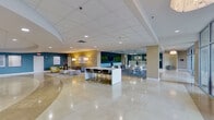 This sophisticated single-floor commercial space is designed to impress, offering a seamless blend of professional functionality and contemporary aesthetics. The grand entrance features polished marble tile flooring and floor-to-ceiling glass walls and doors that flood the interior with natural light while providing views of lush outdoor greenery.

The expansive main lobby serves as the heart of the space, showcasing a gleaming marble floor, recessed lighting, and a drop ceiling with a clean, grid-panel design. Bold teal accent walls punctuate the open layout, adding a vibrant yet professional color palette throughout. Multiple distinct lounge areas are thoughtfully arranged, each furnished with modern sofas, armchairs, area rugs, and accent tables — ideal for informal meetings or relaxed waiting.

A sleek reception area features a prominent desk, a mounted flat-screen display, and warm lighting, offering a welcoming point of contact. A central high-top bar table with bar stools provides a casual collaborative zone. Additional seating alcoves along the corridors offer quiet retreats, while framed artwork and live green wall panels add curated decorative touches.

Glass-partitioned office rooms line portions of the space, maintaining an open feel while providing privacy. The layout flows intuitively from the entry vestibule through the communal zones and into more private areas, making this a well-appointed and versatile professional environment.
