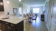 This single-floor residence offers a thoughtfully designed open-concept layout that connects the kitchen, dining, and living areas in a seamless flow. The kitchen features a prominent granite-topped island with dark cabinetry, providing both functionality and an elegant focal point. Hardwood-style flooring extends throughout the main living areas, adding warmth and continuity to the space.

The dining area accommodates a round wooden table surrounded by cushioned upholstered chairs, creating an inviting setting for meals. Just beyond, the living room is bathed in natural light from a large window, offering a bright and airy atmosphere. A large flat-screen television and comfortable seating anchor the living space.

Crown molding runs along the ceilings, adding a refined architectural detail throughout. A decorative potted tree and carefully chosen furnishings contribute to the space's polished, comfortable aesthetic. A glimpse into the bedroom reveals additional light tones and coordinating décor, maintaining a consistent style throughout the residence. The overall ambiance is clean, modern, and welcoming, with attention to detail evident in every corner of this well-appointed single-level home.