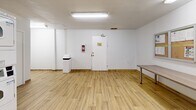 Located at 18-20 Skipton Crt in Toronto, this shared laundry facility offers a clean, efficiently organized space designed for practical, everyday use. The room features light wood-effect flooring throughout, bright white walls, and overhead fluorescent lighting that ensures the space is well-lit at all times. A window provides a degree of natural light to complement the artificial lighting.

The laundry room is equipped with multiple front-loading washing machines and both stacked and top-loading dryers, providing ample capacity for residents. Several utility sinks with faucets are built into the cabinetry along the back wall, adding functional versatility to the space. Visible ventilation ductwork runs along the upper walls, alongside yellow-painted piping, reflecting the utilitarian character of the facility.

A sturdy folding/sorting table is available for residents' convenience, and instructional posters for washer and dryer operation are displayed on the wall for easy reference. A bulletin board is also mounted near the entrance. The room includes a fire extinguisher and a waste bin, prioritizing safety and cleanliness. Two interior doors provide access to adjoining areas, one of which is labeled as a storeroom. The overall ambiance is practical, tidy, and well-maintained, making it a functional communal amenity.