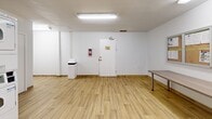 Located at 18-20 Skipton Crt in Toronto, this shared laundry facility offers a clean, efficiently organized space designed for practical, everyday use. The room features light wood-effect flooring throughout, bright white walls, and overhead fluorescent lighting that ensures the space is well-lit at all times. A window provides a degree of natural light to complement the artificial lighting.

The laundry room is equipped with multiple front-loading washing machines and both stacked and top-loading dryers, providing ample capacity for residents. Several utility sinks with faucets are built into the cabinetry along the back wall, adding functional versatility to the space. Visible ventilation ductwork runs along the upper walls, alongside yellow-painted piping, reflecting the utilitarian character of the facility.

A sturdy folding/sorting table is available for residents' convenience, and instructional posters for washer and dryer operation are displayed on the wall for easy reference. A bulletin board is also mounted near the entrance. The room includes a fire extinguisher and a waste bin, prioritizing safety and cleanliness. Two interior doors provide access to adjoining areas, one of which is labeled as a storeroom. The overall ambiance is practical, tidy, and well-maintained, making it a functional communal amenity.
