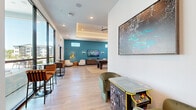 Located at 2500 Conserve Cir in Apopka, FL, this single-floor community lounge and game room offers a generously sized, open-plan recreational space designed for social engagement and entertainment. The interior is defined by a modern aesthetic, featuring light wood-style flooring throughout, recessed lighting, and a tray ceiling with integrated ceiling fans that lend an architectural dimension to the space.

The room is divided into distinct activity zones that flow seamlessly into one another. A dedicated game area anchors the space with a full-size pool table and a long shuffleboard table, complemented by a bold, colorful mural that adds artistic energy to the room. Adjacent to this, a media lounge area features a large flat-screen TV mounted against a striking teal accent wall, accompanied by soft lounge seating and warm pendant lighting. A classic arcade cabinet adds a nostalgic touch near the entrance.

Floor-to-ceiling windows along one side of the room flood the space with abundant natural light and provide views of the outdoor surroundings, while high-top bar seating along the window line creates an inviting spot for casual conversation. Curated black-and-white framed photography adorns the walls, balancing the vibrant mural and colorful accents with a refined, gallery-like quality. The overall ambiance is lively, welcoming, and thoughtfully designed for community interaction.