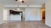 This single-floor space offers an open and airy layout that seamlessly connects the kitchen and living areas. The kitchen is appointed with rich, dark wood cabinetry that provides a striking contrast against the bright white walls and light-toned granite-style countertops. Stainless steel appliances, including a refrigerator and microwave, add a modern touch, while a raised breakfast bar creates a natural boundary between the kitchen and the main living space. Large diagonal-set beige ceramic floor tiles run throughout the open area, lending a sense of spaciousness and visual flow. A ceiling fan is positioned overhead in the living area, contributing to the comfortable and well-ventilated atmosphere. The space benefits from abundant recessed and flush-mount lighting, keeping the interior bright and welcoming. White walls and clean architectural lines give the interior a fresh, neutral palette that adapts easily to a variety of furnishing styles. A set of double doors along one wall provides convenient storage, while a secondary doorway leads further into the unit. The overall ambiance is clean, functional, and move-in ready, with a layout that maximizes usability across every square foot.