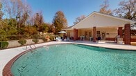 Located at 291 Alabaster Dr in Dothan, Alabama, this single-level outdoor amenity space centers around a generously sized freeform swimming pool with crystal-clear turquoise water and gently sloping entry steps for easy access. The pool is framed by a broad concrete deck finished with a classic brick coping border, providing ample space for lounging and entertaining. A covered pavilion anchored by distinctive brick columns extends from the adjacent building, offering a shaded gathering area outfitted with outdoor dining tables and chairs — an ideal setting for relaxing out of the sun. The building itself features a traditional design with light-colored siding, multiple glass-paned doors, and a gable roof. Additional seating areas are distributed around the pool deck, including cushioned lounge chairs and umbrella-shaded furniture groupings that invite extended outdoor enjoyment. A separate gazebo structure is positioned toward the perimeter, further expanding the outdoor living experience. The entire pool area is enclosed by a wrought-iron fence, ensuring privacy and safety. Mature trees lining the background provide a natural green backdrop and a sense of seclusion, while open skies above fill the space with abundant natural light, creating a bright, resort-like atmosphere throughout the day.