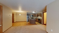 This single-floor space offers a thoughtfully designed open-concept layout that seamlessly connects the kitchen and living areas, creating a comfortable and functional environment. The kitchen is a standout feature, equipped with stainless steel appliances including a refrigerator, microwave, and range, all complemented by rich medium-toned wood cabinetry. A central kitchen island with a granite-style countertop provides additional workspace and serves as a natural gathering point. The kitchen flows into a dedicated dining and living area, where hardwood-style flooring transitions to plush carpeting, adding warmth and texture to the space. A pendant light fixture hangs gracefully over the living area, contributing to the warm, inviting ambiance. The textured ceiling adds a classic architectural touch throughout. Neutral, light-toned walls keep the space bright and versatile, allowing for a variety of interior styles. A secondary hallway off the kitchen leads to additional areas of the unit, hinting at further functional spaces beyond. The entry door, finished in warm wood tones, adds a welcoming touch to the overall design. This space is ideal for those seeking a practical yet comfortable single-level layout.