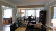 Located at 5500 Wissahickon Ave in the Germantown neighborhood of Philadelphia, PA, this single-floor residence offers a well-appointed and cohesive living space designed for both comfort and style. The open-concept layout seamlessly connects the living and dining areas, creating a spacious and inviting atmosphere. Crown molding and a subtle beam detail add architectural character to the neutral-toned interior.

The dining area is a standout feature, anchored by a graceful bay window that floods the space with natural light and provides an airy, open feel. Sheer patterned curtains frame the window elegantly, complementing the classic round dining table and dark upholstered chairs arranged beneath it.

Adjacent to the dining space, the living area is anchored by a tufted dark sofa adorned with an eclectic mix of decorative pillows in teal, gold, and geometric patterns, adding warmth and personality to the room. A dark wood armoire provides substantial storage while contributing to the room's rich, layered aesthetic. Hardwood flooring runs throughout, tying the spaces together with a sense of continuity and timeless appeal. The overall ambiance is sophisticated yet welcoming, blending classic architectural details with thoughtfully curated furnishings.