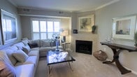 Located at 10212 Cascade Falls Ct, this single-floor home offers a thoughtfully designed living space with a warm and refined aesthetic. The living room serves as the heart of the home, centered around a classic tile-surround fireplace framed by a light-toned mantle, above which decorative artwork adds a polished touch. Soft, neutral-toned carpeting runs throughout the space, contributing to the room's comfortable and understated elegance. Plantation shutters on the large windows provide both privacy and excellent light control, allowing natural light to filter in and brighten the interior. The walls are finished in a soothing gray-beige palette that complements the neutral furnishings and décor. Crown molding traces the ceiling perimeter, adding an architectural detail that elevates the overall sophistication of the space. A sleek glass-top coffee table and a generously sized sectional sofa anchor the seating area, creating an inviting setting for relaxation or entertaining. The single-floor layout ensures seamless flow and accessibility throughout. Overall, the space exudes a calm, well-appointed ambiance that balances comfort with classic style.