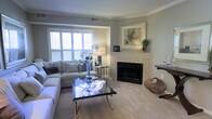 Located at 10212 Cascade Falls Ct, this single-floor home offers a thoughtfully designed living space with a warm and refined aesthetic. The living room serves as the heart of the home, centered around a classic tile-surround fireplace framed by a light-toned mantle, above which decorative artwork adds a polished touch. Soft, neutral-toned carpeting runs throughout the space, contributing to the room's comfortable and understated elegance. Plantation shutters on the large windows provide both privacy and excellent light control, allowing natural light to filter in and brighten the interior. The walls are finished in a soothing gray-beige palette that complements the neutral furnishings and décor. Crown molding traces the ceiling perimeter, adding an architectural detail that elevates the overall sophistication of the space. A sleek glass-top coffee table and a generously sized sectional sofa anchor the seating area, creating an inviting setting for relaxation or entertaining. The single-floor layout ensures seamless flow and accessibility throughout. Overall, the space exudes a calm, well-appointed ambiance that balances comfort with classic style.