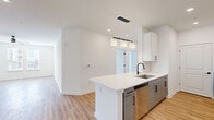 This single-floor residence showcases a clean, modern design with a cohesive neutral palette throughout. The kitchen is a standout feature, equipped with two-tone cabinetry — white upper cabinets paired with gray lower cabinets — accented by matte black hardware. White quartz countertops, a subway tile backsplash, and stainless steel appliances including a refrigerator, electric range, microwave, and dishwasher complete the space. A peninsula island with an undermount sink provides additional prep space and opens toward the living area, which is illuminated by pendant lighting and a set of glass-paneled doors that invite natural light. Light wood-look flooring runs throughout the main living areas, creating a warm, unified feel. The bedrooms are carpeted for comfort, each featuring built-in closets with bifold doors and windows that bring in natural light. The bathroom is finished with large-format gray tile in the tub surround, a gray vanity with white countertop, wood-look flooring, and matte black fixtures. A dedicated entryway area with louvered closet doors and additional storage adds practicality to the overall layout.
