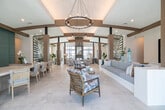 Located at 17174 Curry Preserve Dr in Punta Gorda, Florida, this generously sized single-floor community clubhouse serves as the social hub of the Canopy development. The space is defined by a bright, open design characterized by large-format travertine tile flooring, soaring ceilings with exposed wood beam accents, and an abundance of natural light flowing through floor-to-ceiling black-framed windows and glass doors.

Upon entry, wide, well-appointed hallways feature built-in wood shelving and curated wall art, guiding visitors into the expansive main lobby. The reception area is anchored by a bold teal paneled accent wall bearing the "Canopy" brand, surrounded by comfortable seating and elegant chandelier lighting. The living room area extends into a generously scaled lounge featuring a billiard table, built-in banquette seating with textured wall paneling, and a large wall-mounted television.

A standout stone-clad fireplace, framed by floor-to-ceiling glass walls overlooking a sunny outdoor terrace with palm views, serves as a dramatic focal point. The fully equipped kitchen offers wood cabinetry, white quartz countertops, a sage green tile backsplash, and multiple windows capturing outdoor Florida scenery. A glass-enclosed conference room with sliding barn-style doors, a long wooden table, and rope-style chandeliers completes this well-rounded community facility.