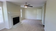 This single-story space centers around a comfortable living area finished with neutral wall-to-wall carpeting that lends a warm, understated feel throughout. The room is anchored by a tile-surround fireplace with a classic white mantel and a black firebox insert, serving as a natural focal point. A ceiling fan with a built-in light fixture is centrally mounted overhead, providing both air circulation and ambient lighting. Natural light filters into the space through a window positioned near the fireplace wall, brightening the neutral, light-toned walls. The layout is open and unobstructed, offering flexible furniture placement options. A standard interior door on the far wall provides access to an adjoining area or room. The overall ambiance is clean, simple, and move-in ready, with a neutral color palette throughout that suits a wide range of interior styles. The combination of the fireplace, carpeted flooring, and ceiling fan creates a functional and inviting living environment well-suited for everyday comfort.
