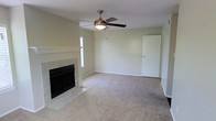 This single-story space centers around a comfortable living area finished with neutral wall-to-wall carpeting that lends a warm, understated feel throughout. The room is anchored by a tile-surround fireplace with a classic white mantel and a black firebox insert, serving as a natural focal point. A ceiling fan with a built-in light fixture is centrally mounted overhead, providing both air circulation and ambient lighting. Natural light filters into the space through a window positioned near the fireplace wall, brightening the neutral, light-toned walls. The layout is open and unobstructed, offering flexible furniture placement options. A standard interior door on the far wall provides access to an adjoining area or room. The overall ambiance is clean, simple, and move-in ready, with a neutral color palette throughout that suits a wide range of interior styles. The combination of the fireplace, carpeted flooring, and ceiling fan creates a functional and inviting living environment well-suited for everyday comfort.