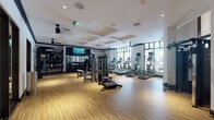 Located at 600 N Pine Island Rd in Plantation, FL, this well-appointed fitness center offers a comprehensive and modern workout environment across a single, open floor. The space is defined by warm hardwood-style flooring, crisp white walls with bold black trim accents, and a coffered ceiling outfitted with recessed lighting and ceiling fans that enhance both style and comfort.

Large floor-to-ceiling windows line one wall, flooding the cardio zone with natural light and offering views of the lush outdoor surroundings. This area is equipped with a row of treadmills, elliptical trainers, and stair climbers from commercial-grade brands including Star Trac and Stairmaster, each featuring individual display screens.

The free weight section features a full rack of dumbbells, weight benches, and wall-mounted mirrors, creating an ideal environment for strength training. A dedicated functional training area includes a TRX suspension system, resistance bands, stability balls, and a wall-mounted rig for varied bodyweight and resistance exercises.

A cable machine and additional strength equipment occupy the central floor space, offering versatile training options. Glass-paneled black-framed doors connect distinct zones within the facility, maintaining an open yet organized layout. A dramatic black spiral staircase serves as a striking architectural focal point, adding a sculptural element to the overall aesthetic. The facility combines functionality with refined design throughout.