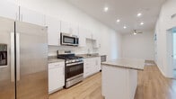 This well-appointed single-floor unit showcases a clean, contemporary design throughout, with crisp white walls and light wood-tone flooring creating a bright and cohesive aesthetic. The kitchen is a standout feature, offering white shaker-style cabinetry with sleek bar-pull hardware, granite countertops, and a full suite of stainless steel appliances including a gas range, over-the-range microwave, side-by-side refrigerator with ice and water dispenser, and a dishwasher. A kitchen island provides additional counter space and storage, enhancing the functionality of the layout. The living area extends naturally from the kitchen, maintaining the light wood flooring and featuring a ceiling fan overhead. A bedroom offers soft carpeted flooring and benefits from generous natural light through a pair of large windows, creating a calm and comfortable retreat. The bathroom is finished with matching white cabinetry, granite countertops, a modern single-basin undermount sink with a contemporary faucet, and a full shower-over-tub combination. Recessed lighting throughout adds a polished finishing touch, while the consistent use of white tones and modern materials gives the entire space a fresh, move-in-ready feel.