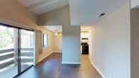 This single-story space showcases an interesting architectural design defined by varying ceiling heights that create visual depth and dimension throughout the interior. The living area features a vaulted ceiling with white-painted wood plank paneling and exposed beams, adding a warm, textured contrast to the smooth textured ceilings found in adjacent areas. Dark hardwood-style flooring runs throughout, providing a grounding element against the neutral, light taupe wall tones.

The open layout connects the main living space to a secondary area, which is equipped with a ceiling fan and a large window that invites natural light. Sliding glass doors along one wall further enhance the sense of openness and provide access to the exterior. A partial wall divides the space, creating a natural transition to the kitchen, which features light wood-style flooring, white cabinetry, and black appliances including a range with overhead ventilation. Recessed lighting illuminates the kitchen area. The overall ambiance is clean and functional, with a neutral palette that offers a versatile canvas throughout.