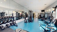 Located on Navarre Parkway in Navarre, Florida, this single-floor fitness center offers a comprehensive workout facility divided into two dedicated exercise rooms connected by a hallway. The space is thoughtfully designed with a bright, open feel, thanks to numerous large windows that flood the interior with natural light and provide views of the surrounding outdoor pool area and landscaping.

The first exercise room is dedicated to strength training and features a variety of cable machines, multi-station weight equipment, adjustable benches, and free weights, all set against full wall-to-wall mirrors that enhance the sense of space and allow users to monitor their form. The vivid blue flooring adds a clean, energetic aesthetic throughout.

The second exercise room focuses on cardiovascular training and is outfitted with multiple treadmills, stationary bikes, ellipticals, and a stair climber. A wall-mounted flat-screen television provides entertainment during workouts. Floor-to-ceiling mirrors line one wall, further amplifying the light and spacious atmosphere.

Recessed ceiling lighting supplements the natural light throughout both rooms, ensuring well-lit conditions at any time of day. The facility's clean white walls, durable sport flooring, and modern equipment selections create a professional and motivating environment suited for a wide range of fitness routines.