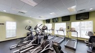 This fitness center offers a dedicated space for cardio-focused workouts, thoughtfully arranged to maximize both functionality and comfort. The room features sage-green accent walls paired with a drop-tile ceiling fitted with recessed and panel lighting, creating a clean, well-lit environment conducive to exercise.

The cardio equipment lineup includes multiple elliptical trainers positioned along the left side of the room, complemented by a stationary bike and a row of treadmills extending toward the far wall. This variety of machines accommodates a range of workout preferences and fitness levels.

A bank of large windows along the back wall allows generous natural light to fill the space while offering exterior views, enhancing the overall workout experience. Multiple flat-screen televisions are mounted at eye level along the walls, providing entertainment options during exercise sessions.

The flooring transitions from a standard surface beneath the ellipticals to a darker tread beneath the treadmills, adding a practical design element. A wall clock and informational signage complete the utilitarian yet inviting atmosphere of this well-maintained fitness facility.
