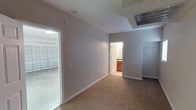 This single-floor space presents a clean, functional layout designed for practical use. The main room is finished with wall-to-wall carpeting in a neutral tan tone, complemented by light gray walls and white baseboards that give the interior a fresh, understated appearance. A large HVAC vent is mounted in the ceiling, ensuring climate control throughout the space. Electrical outlets are positioned along the walls for convenient access.

A doorway leads directly into an attached bathroom, which features warm wood-toned cabinetry, a sink, and tile flooring, providing essential amenities in a compact and efficient design. On the opposite side of the main room, a wide doorway opens into an attached garage, which has a concrete floor and a roll-up garage door, adding significant utility and storage potential to the overall space.

The layout is straightforward and open, with no unnecessary partitions, making the space highly adaptable for a variety of uses. The neutral color palette throughout contributes to a clean, move-in-ready atmosphere, while the combination of carpeted living area, private bathroom, and direct garage access makes this a notably versatile and self-contained unit.