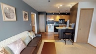 This single-floor unit offers a thoughtfully designed layout that blends functionality with a distinct, modern aesthetic. The open-concept living and dining area flows seamlessly into the kitchen, creating a connected and sociable space. Bold charcoal-gray accent walls contrast with warm hardwood-style flooring throughout the main living area, giving the space a contemporary feel. A track lighting system illuminates both the living space and kitchen, ensuring bright, flexible lighting throughout.

The kitchen is well-equipped, featuring warm honey-toned wood cabinetry, laminate countertops, a full suite of appliances including a refrigerator, electric range, and dishwasher, and a compact breakfast bar that provides additional seating and counter space. A decorative "eat" wall accent adds a charming, personalized touch.

The two bedrooms are comfortably sized and feature plush blue carpeting, ample natural light from windows with blinds, and generous closet space. One bedroom is accented with a teal feature wall, adding a pop of color and personality.

The bathroom is finished with large-format tile flooring, a light wood vanity with an integrated sink, a wall-mounted mirror, and overhead lighting, providing a clean and functional space. Overall, the unit presents a cohesive and inviting interior with practical storage and comfortable living spaces throughout.