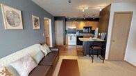 This single-floor unit offers a thoughtfully designed layout that blends functionality with a distinct, modern aesthetic. The open-concept living and dining area flows seamlessly into the kitchen, creating a connected and sociable space. Bold charcoal-gray accent walls contrast with warm hardwood-style flooring throughout the main living area, giving the space a contemporary feel. A track lighting system illuminates both the living space and kitchen, ensuring bright, flexible lighting throughout.

The kitchen is well-equipped, featuring warm honey-toned wood cabinetry, laminate countertops, a full suite of appliances including a refrigerator, electric range, and dishwasher, and a compact breakfast bar that provides additional seating and counter space. A decorative "eat" wall accent adds a charming, personalized touch.

The two bedrooms are comfortably sized and feature plush blue carpeting, ample natural light from windows with blinds, and generous closet space. One bedroom is accented with a teal feature wall, adding a pop of color and personality.

The bathroom is finished with large-format tile flooring, a light wood vanity with an integrated sink, a wall-mounted mirror, and overhead lighting, providing a clean and functional space. Overall, the unit presents a cohesive and inviting interior with practical storage and comfortable living spaces throughout.