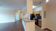 This single-story space showcases an open-concept layout that seamlessly connects the kitchen and living areas. The kitchen is a functional centerpiece, featuring warm honey-toned wood cabinetry that provides ample storage, granite-patterned countertops, and a full suite of black appliances, including a refrigerator, electric range, and built-in microwave. A peninsula with a dual-basin sink and matching countertop extends outward, creating a natural division between the kitchen and the adjoining living space while offering additional prep and seating space.

The flooring throughout is dark hardwood-style, adding warmth and visual continuity across the open floor plan. Structural columns with a textured finish are incorporated into the design, framing the kitchen area and adding architectural character. The ceilings feature a textured finish and are fitted with recessed and flush-mount lighting to ensure well-lit interiors.

A hallway visible from the main living area leads to additional rooms, with a set of bifold doors hinting at closet or utility storage. The overall palette is neutral, with beige and cream tones on the walls complementing the darker flooring, creating a balanced and inviting atmosphere throughout the space.