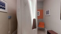 Located at 125 N Mill Rd in Vineyard, Utah, this single-room tanning space is a compact yet thoughtfully designed facility. At its centerpiece stands a sleek Luxura tanning unit, its smooth white casing and built-in accent lighting making it an immediate focal point. The room's interior design is characterized by a bold two-tone color scheme — vibrant orange upper walls paired with soft gray lower sections — creating an energetic and welcoming atmosphere. Framed geometric and patterned artwork adorns the walls, adding a stylish, contemporary touch throughout the space. A modern orange lounge chair provides a comfortable seating area for clients, complementing the room's color palette. Light-toned ceramic tile flooring runs throughout, contributing to the clean and polished aesthetic. A wooden ladder-style shelf near the entry door holds sanitizing supplies, wicker storage baskets, and instructional signage, ensuring the space remains organized and hygienic. A decorative mirror with an ornate frame, a low storage bench, and a stainless steel step trash can add functional elegance. A coat hook rail and an air purifier further enhance comfort and practicality. The white paneled entry door with brushed nickel hardware completes the tidy, professional finish of this well-appointed tanning room.