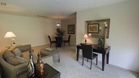 This single-floor residence offers a thoughtfully designed open-concept layout that seamlessly connects the living and dining areas, creating a welcoming and functional environment. The living area features plush wall-to-wall carpeting in a neutral tone, providing a soft, comfortable foundation that complements the warm, earthy color palette used throughout the space. A stylishly furnished seating area anchors the room, accented by decorative pieces that add character and depth. Adjacent to the living area, a dedicated desk or console station is arranged with a dark-finished writing desk, a cushioned chair, and a large ornate mirror that reflects light and enhances the sense of space. A warm table lamp adds an inviting glow to this corner. The dining area is defined by a classic round table surrounded by dark upholstered chairs, illuminated by an elegant pendant light fixture that hangs from a textured ceiling. A tall potted plant and framed wall art contribute to the polished, refined atmosphere of the dining space. Throughout the residence, neutral wall tones and consistent flooring create a cohesive aesthetic, while the overall layout promotes an easy flow between functional areas, making this space both practical and visually appealing.